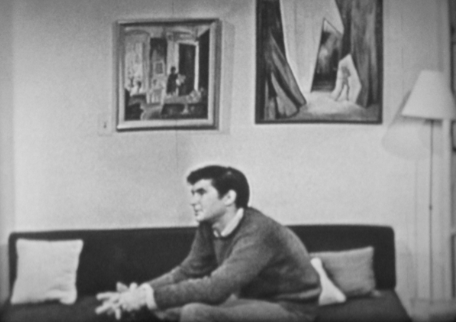 A man sits on a couch in a living room with abstract paintings, evoking themes of presence and Urdu poetry.