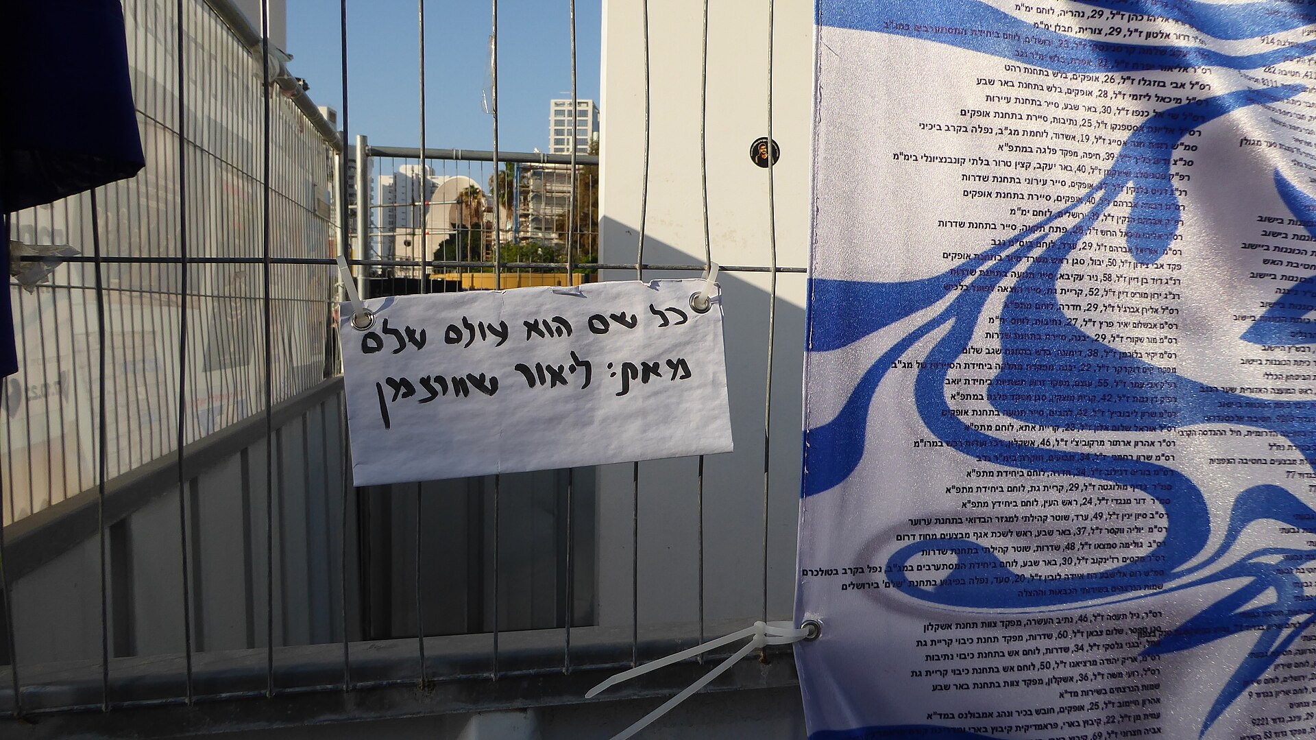 A protest sign with Hebrew writing is displayed on a metal fence, reflecting themes of political activism.